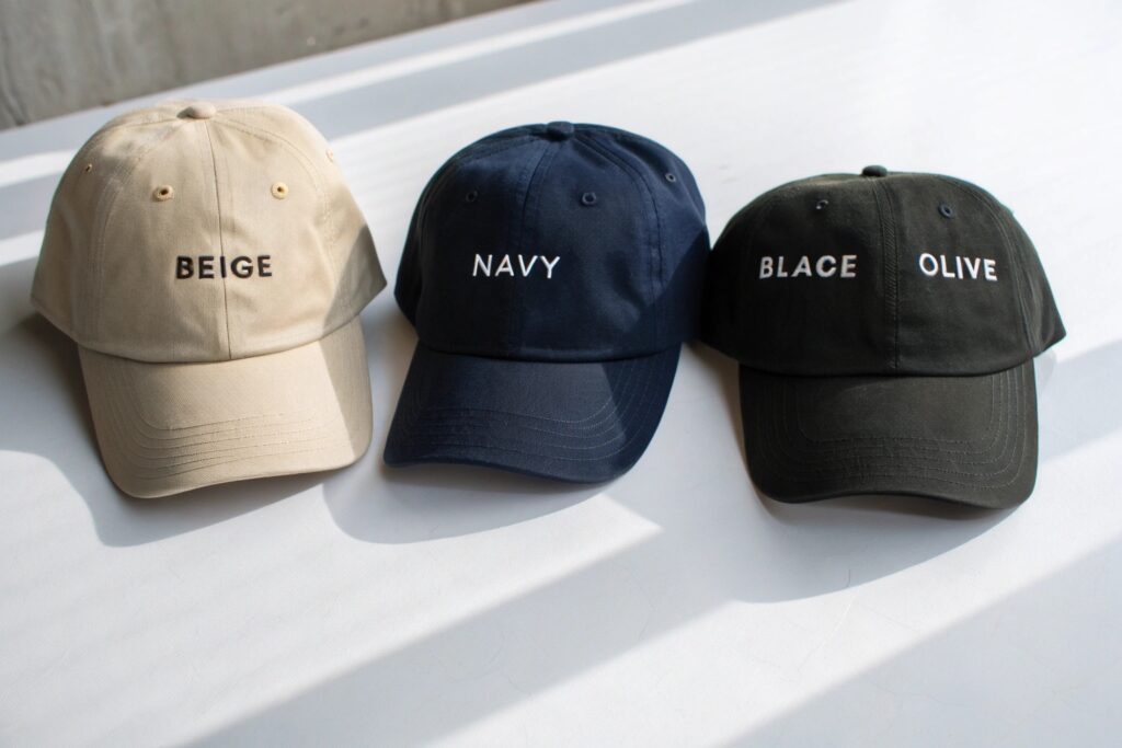 Top-down product photography of four custom embroidered dad hats in neutral tones – beige, navy, black, and olive. Perfect for showcasing unstructured cap colorways, casual branding, and minimalist embroidered typography. Ideal for wholesale headwear line sheets or e-commerce product listings.