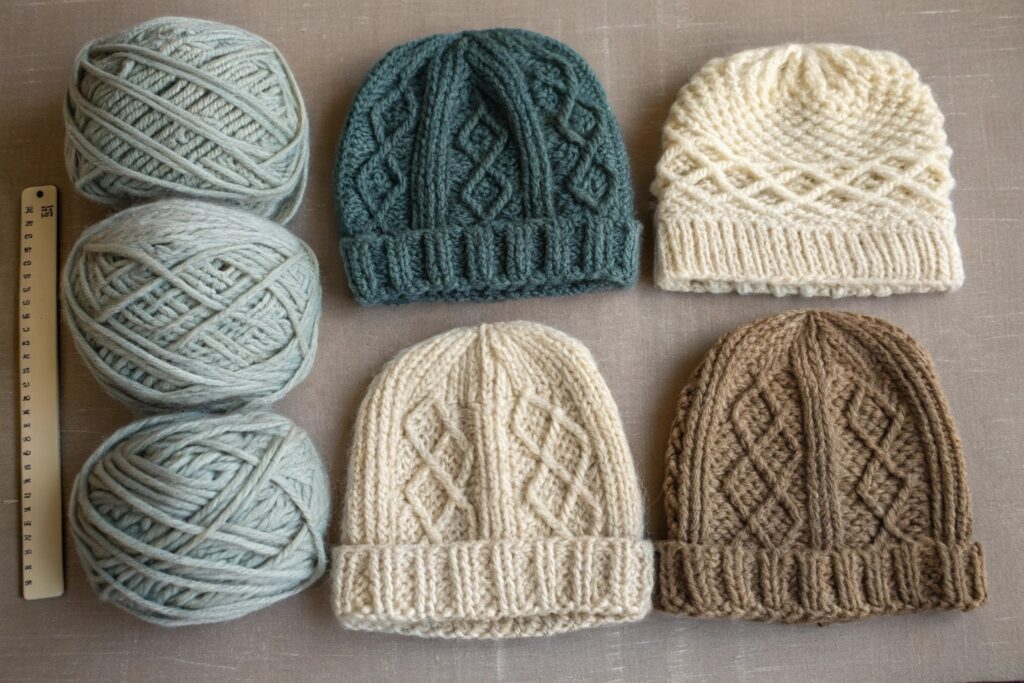 A flat lay of four intricately hand-knitted beanies in different earth tones, arranged next to three balls of yarn and a ruler, showcasing texture, size, and craftsmanship — ideal for DIY enthusiasts and wool product showcases