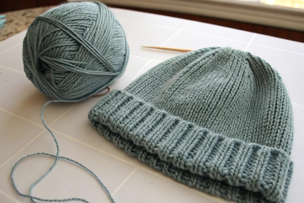 A finished knitted hat placed beside a matching ball of yarn, highlighting the soft texture and uniform stitching of the hand-knit design