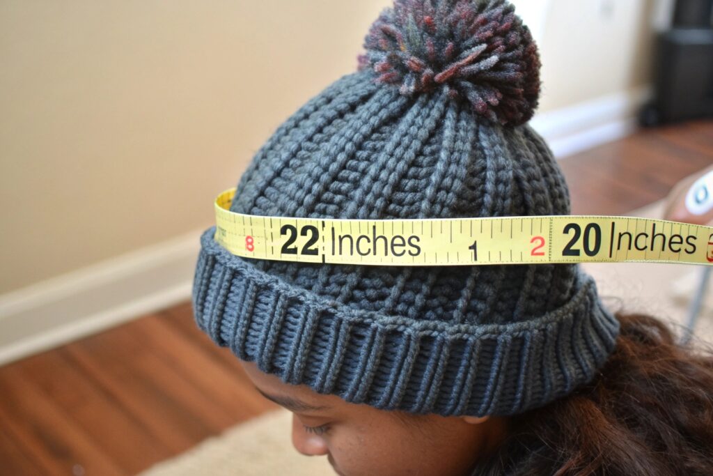 A person wearing a gray knit hat with a pom-pom on top, with a yellow measuring tape wrapped around their head