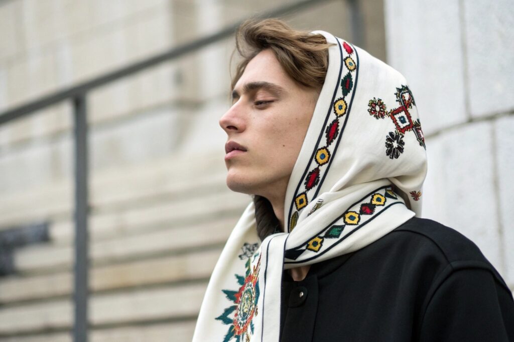 A person with light brown hair wearing a white headscarf with colorful embroidered patterns and a black jacket, eyes closed